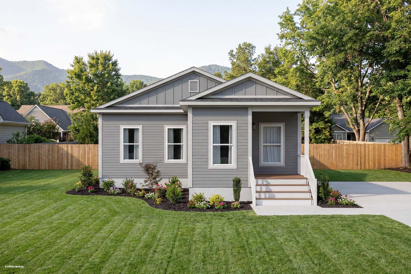 Small gray modular home with white trim, front porch, neat lawn, flowerbeds, and trees in the background.