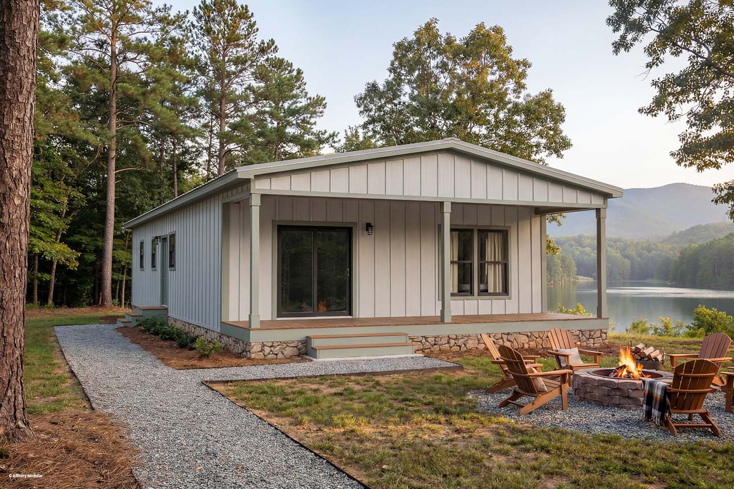 A modern modular home on the lakefront with a fire pit and Adirondack chairs, surrounded by trees and mountains.
