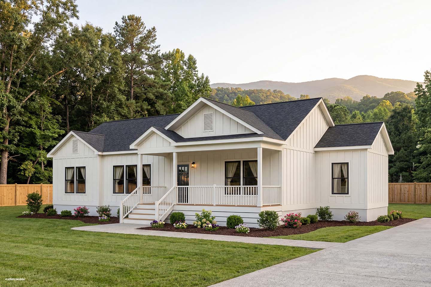 A modern white farmhouse modular home with a front porch, surrounded by landscaping and trees, with mountains in the background.